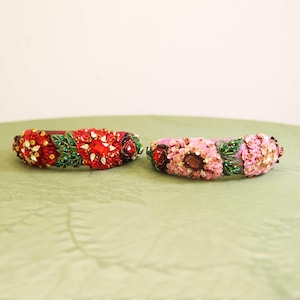 May include: Two ornate headbands. One is red with gemstones, gold accents, and green leaves. The other is pink with flowers and a large brown gem. Both are embellished with beads and sequins, perfect for special occasions.
