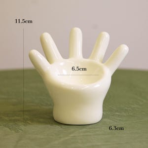 以下が含まれることがあります： クリーム色のセラミック製の手の形をした小皿。高さ11.5cm、幅6.5cm、奥行き6.3cm。手のひらに浅いボウルがあり、小物を入れるように設計されています。指は完全に伸びています。