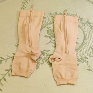 May include: A pair of beige compression stockings with zippers running down the front. The stockings are knee-high and have a foot opening. The zippers are tan and have a small pull tab at the top. The stockings are made of a soft, stretchy material.
