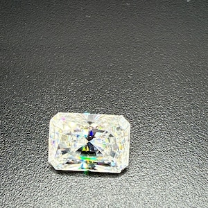 May include: A radiant-cut diamond with a rectangular shape, showcasing brilliant facets. The gemstone sparkles with a spectrum of colors, reflecting light. The diamond is set against a dark, textured background.