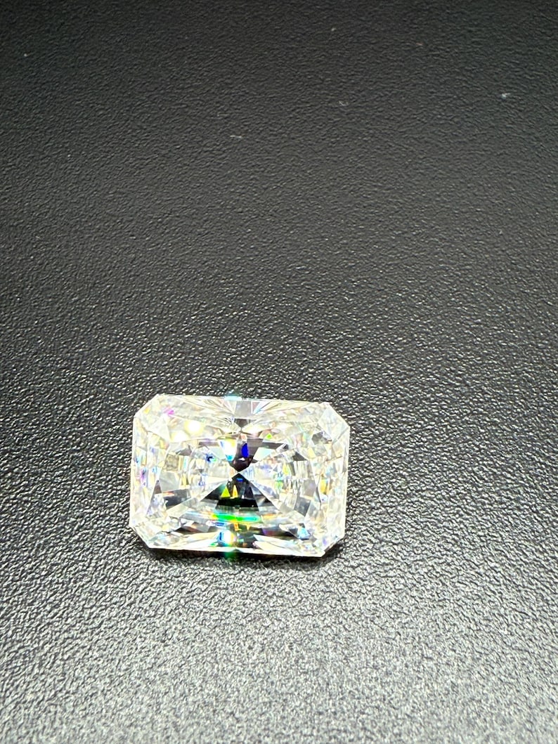 May include: A radiant-cut diamond with a rectangular shape, showcasing brilliant facets. The gemstone sparkles with a spectrum of colors, reflecting light. The diamond is set against a dark, textured background.