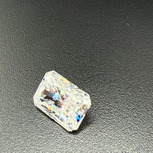 May include: A radiant-cut diamond with a rectangular shape. The gemstone is clear and reflects a spectrum of colors, including blue, green, and yellow. The diamond is set against a dark background.
