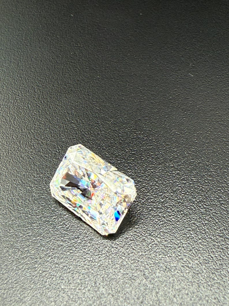 May include: A radiant-cut diamond with a rectangular shape. The gemstone is clear and reflects a spectrum of colors, including blue, green, and yellow. The diamond is set against a dark background.