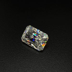 Può includere: Un diamante a taglio radiante rettangolare, che mostra un gioco di colori nelle sue sfaccettature. La gemma è posta su uno sfondo scuro, evidenziando la sua brillantezza e chiarezza. Le sfaccettature del diamante riflettono la luce, creando un'esibizione accattivante.