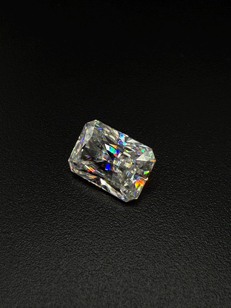 May include: A radiant-cut diamond with a rectangular shape, displaying a spectrum of colors within its facets. The gemstone is set against a dark background, highlighting its brilliance and clarity. The diamond's facets catch and reflect light, creating a dazzling display.