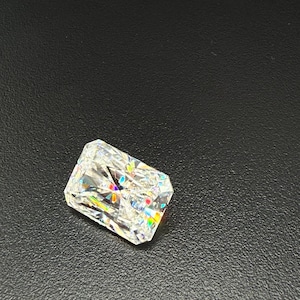 May include: A radiant-cut diamond with a rectangular shape. The gemstone is clear and reflects a spectrum of colors, including red, yellow, and blue. The diamond is set against a dark, textured background.