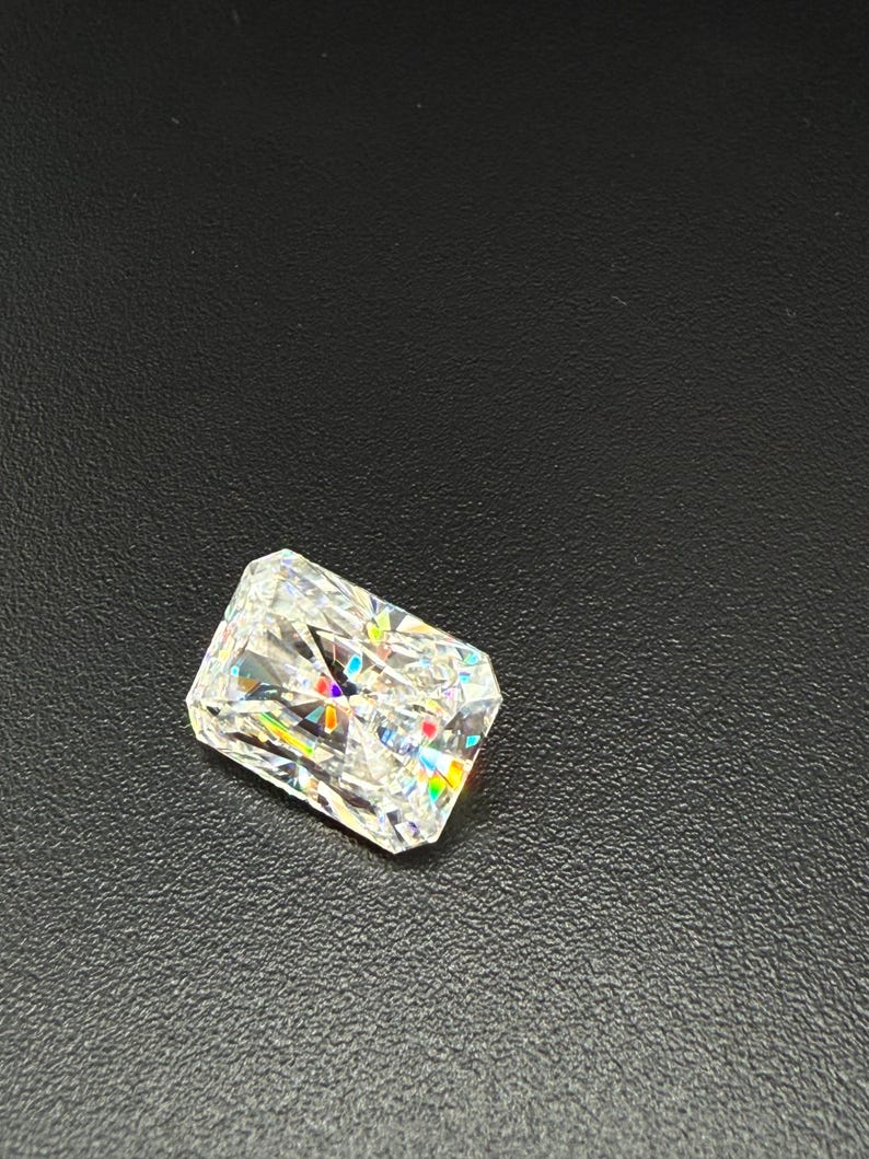 May include: A radiant-cut diamond with a rectangular shape. The gemstone is clear and reflects a spectrum of colors, including red, yellow, and blue. The diamond is set against a dark, textured background.
