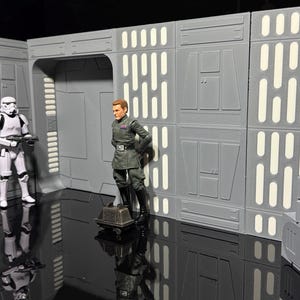 Może przedstawiać: Zestaw diorama Star Wars z figurką szturmowca i figurką oficera imperialnego. Zestaw ma szare ściany z białymi akcentami i odblaskową czarną powierzchnią.