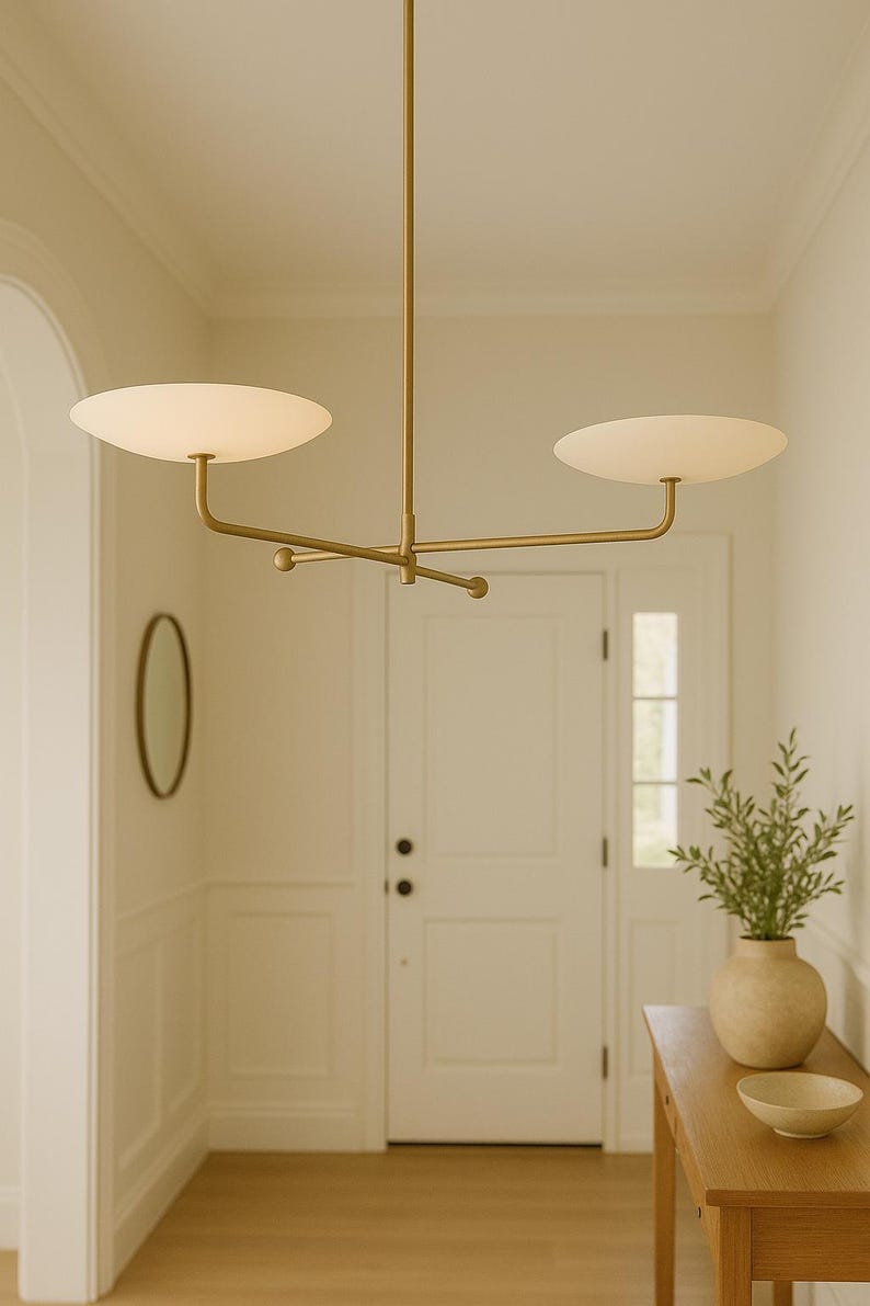Modern brass linear chandelier with two dish-shaped shades hanging above a minimalist gray and white bedroom interior
Handmade brass pendant light fixture with dual disc shades in a modern bedroom with neutral décor
Luxury brushed brass two-arm linear chandelier installed above a bed in a contemporary minimalist room
Mid century modern brass ceiling light with two flat dome shades illuminating a cozy bedroom interior