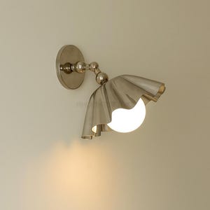 K&ouml;nnte beinhalten: Eine silberfarbene Wandleuchte mit einzigartigem Design. Sie hat eine kreisf&ouml;rmige Basis, einen gebogenen Arm mit Kugelgelenken und einen ger&uuml;schten Schirm, der ein kugelf&ouml;rmiges, leuchtendes Licht umgibt. Die Leuchte ist an einer neutralfarbenen Wand befestigt.