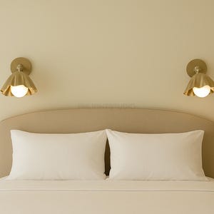 K&ouml;nnte beinhalten: Ein Schlafzimmer mit einem Bett mit wei&szlig;er Bettw&auml;sche und Kissen. Zwei goldfarbene Wandleuchten mit bl&uuml;tenblattf&ouml;rmigem Design und einer kugelf&ouml;rmigen Gl&uuml;hbirne sind &uuml;ber dem Bett angebracht und sorgen f&uuml;r sanfte Beleuchtung.