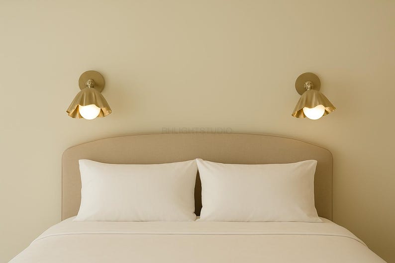 K&ouml;nnte beinhalten: Ein Schlafzimmer mit einem Bett mit wei&szlig;er Bettw&auml;sche und Kissen. Zwei goldfarbene Wandleuchten mit bl&uuml;tenblattf&ouml;rmigem Design und einer kugelf&ouml;rmigen Gl&uuml;hbirne sind &uuml;ber dem Bett angebracht und sorgen f&uuml;r sanfte Beleuchtung.
