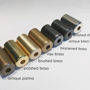 K&ouml;nnte beinhalten: Eine Sammlung zylindrischer Metallobjekte in verschiedenen Ausf&uuml;hrungen, darunter poliertes Chrom, Antikschwarz und geb&uuml;rstetes Messing. Das Bild zeigt eine Reihe von Farben und Texturen, wobei Etiketten jede Ausf&uuml;hrung kennzeichnen. Die Objekte sind in einer Reihe angeordnet.