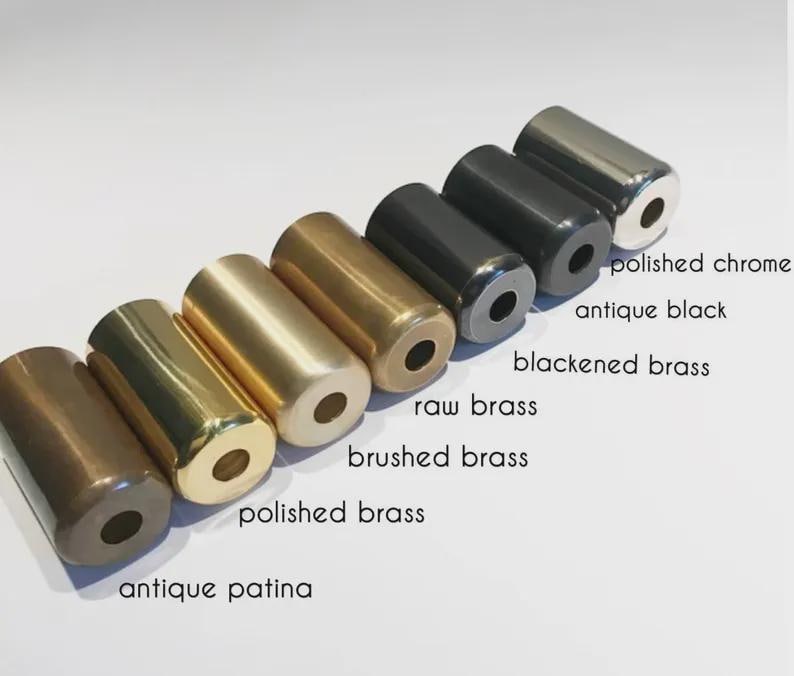 K&ouml;nnte beinhalten: Eine Sammlung zylindrischer Metallobjekte in verschiedenen Ausf&uuml;hrungen, darunter poliertes Chrom, Antikschwarz und geb&uuml;rstetes Messing. Das Bild zeigt eine Reihe von Farben und Texturen, wobei Etiketten jede Ausf&uuml;hrung kennzeichnen. Die Objekte sind in einer Reihe angeordnet.