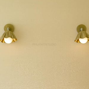K&ouml;nnte beinhalten: Zwei Wandleuchten mit goldener Oberfl&auml;che, jede mit einem gewellten Schirm und einem runden, leuchtenden Licht. Die Leuchten sind an einer hellen Wand montiert und schaffen ein warmes und einladendes Ambiente.