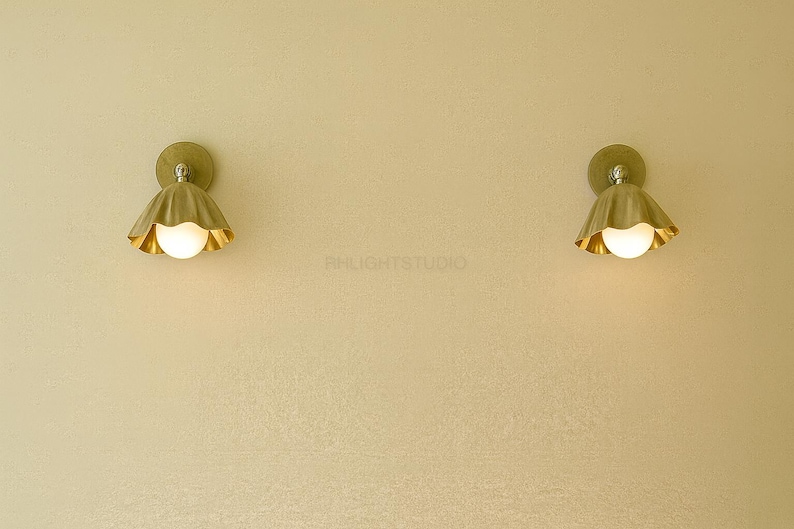 K&ouml;nnte beinhalten: Zwei Wandleuchten mit goldener Oberfl&auml;che, jede mit einem gewellten Schirm und einem runden, leuchtenden Licht. Die Leuchten sind an einer hellen Wand montiert und schaffen ein warmes und einladendes Ambiente.