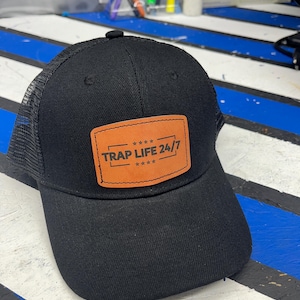 May include: Black trucker hat with a mesh back and a curved brim. The front features a brown leather patch with the text "TRAP LIFE 24/7" in black lettering and four stars.