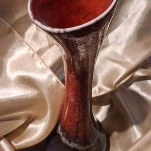 May include: A tall ceramic vase with a flared, wide opening. The interior and upper neck feature a glossy, mottled red glaze that transitions into a dark, solid black base. The surface has a smooth, reflective finish with subtle colour variations.