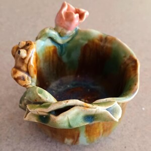 Puede incluir: Un cuenco de cerámica con un diseño de rana y flor. El cuenco es principalmente verde, marrón y naranja, con una figura de rana en el lateral y una flor rosa en la parte superior. El cuenco tiene una forma orgánica única.
