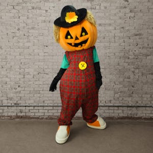Könnte beinhalten: Ein Maskottchenkostüm mit einem großen orangefarbenen Kürbiskopf mit schwarzem Jack-o'-Lantern-Gesicht, das einen schwarzen Hut mit einem gelben Blatt trägt. Der Körper ist ein rot-grüner karierter Overall mit einem gelben Knopf, türkisfarbenen Ärmeln, schwarzen Handschuhen und weißen Schuhen.