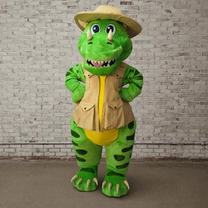 May include: A green dinosaur costume with a tan safari-style vest and hat. The costume has large, friendly eyes, a toothy grin, and clawed feet. The costume is designed to be worn by a person.