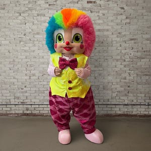 May include: A full-body clown costume with a rainbow wig, a yellow vest, and pink pants. The costume features a large, smiling face with green eyes, a red nose, and a pink bow tie. The clown is wearing pink shoes.