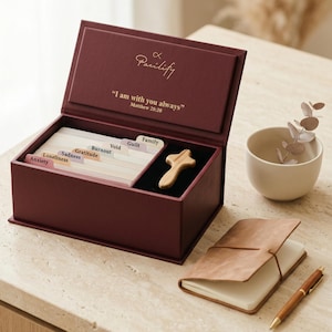 K&ouml;nnte beinhalten: Eine tiefburgunderrote rechteckige Box mit Klappdeckel, die den Text Pacilify und I am with you always Matthew 28:20 tr&auml;gt. Im Inneren befinden sich Registerkarten mit den Beschriftungen Anxiety, Loneliness, Sadness, Gratitude, Burnout, Void, Guilt und Family sowie ein Holzkreuz.