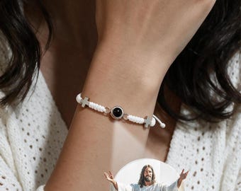 Könnte beinhalten: Ein weißes Armband aus geflochtener Kordel mit zwei silberfarbenen Kreuzanhängern und einem zentralen runden Anhänger, der ein Bild von Jesus mit ausgebreiteten Armen projiziert, darunter der Text "I am with you always".