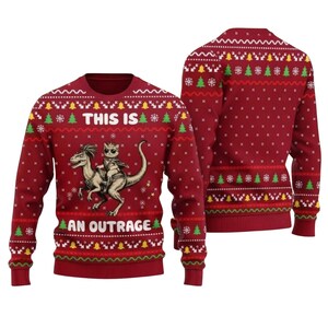 May include: A red long-sleeve crewneck jumper featuring a repeating white and green holiday pattern of snowflakes, trees, and bells. The front displays a graphic of a cat riding a dinosaur with the text THIS IS AN OUTRAGE in white letters.