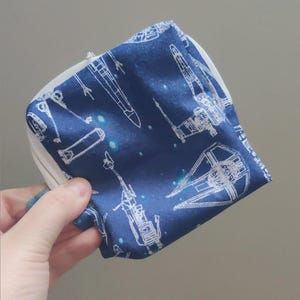May include: A small, square, zippered pouch with a navy blue background and white line drawings of spaceships. The pouch is held in a hand, and the zipper is white. The design includes various spacecraft and a starry sky.