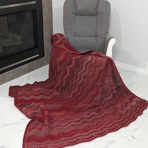May include: A handmade, red and burgundy crochet blanket with a wavy pattern is draped over a grey armchair. The blanket is made of soft yarn and has a textured appearance. The blanket is a cosy home decor item.