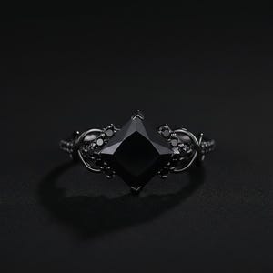 May include: A black ring with a large, square-cut black gemstone as the centerpiece. The ring has an intricate, dark metal band with smaller black stones and decorative details. The overall design is gothic and elegant.