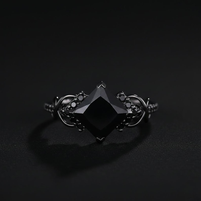 May include: A black ring with a large, square-cut black gemstone as the centerpiece. The ring has an intricate, dark metal band with smaller black stones and decorative details. The overall design is gothic and elegant.