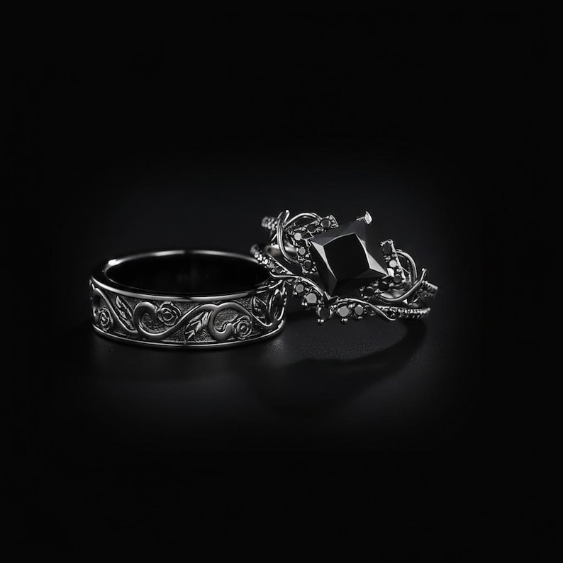 May include: Two black rings. One ring features a floral design, while the other has a large, square black gemstone and smaller black stones. The rings are set against a black background.