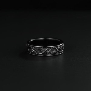 May include: A dark, silver-toned ring with an intricate floral design. The ring features a repeating pattern of roses and leaves around the band. The ring is set against a dark background, highlighting the detailed craftsmanship.