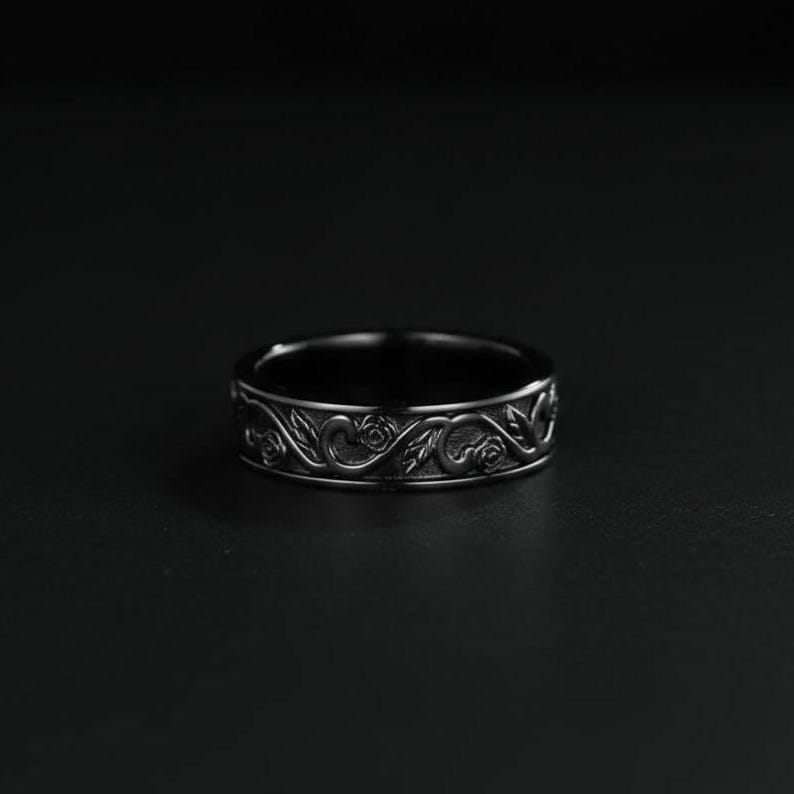 May include: A dark, silver-toned ring with an intricate floral design. The ring features a repeating pattern of roses and leaves around the band. The ring is set against a dark background, highlighting the detailed craftsmanship.