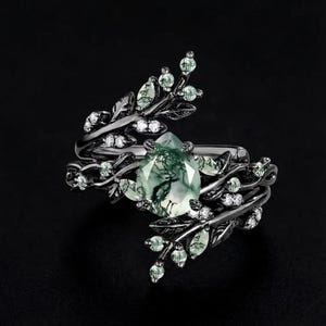 Puede incluir: Un anillo ornamentado con una piedra preciosa verde en forma de pera como punto focal. El diseño del anillo incorpora delicados detalles en forma de hojas con pequeñas gemas transparentes y una banda de metal oscuro. La estética general es de inspiración natural y elegante.