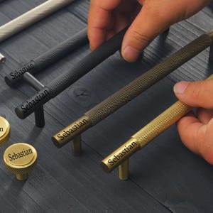 Op de afbeelding: Een collectie metalen kastbeslag inclusief lange cilindrische staafgrepen en ronde knoppen met een getextureerd gekarteld oppervlak. Het beslag is verkrijgbaar in diverse metalen afwerkingen waaronder zilver, matzwart, messing en goud, elk voorzien van het label Sebastian.