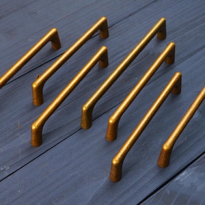 May include: A set of six gold-toned metal cabinet pulls arranged in two rows. Each handle features a smooth, arched design with two mounting points at the ends, showcasing a consistent metallic finish and a curved, elongated bar structure.