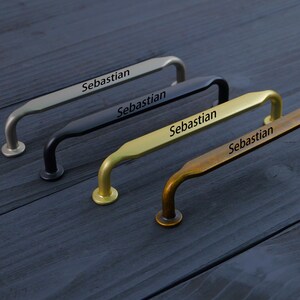 May include: Four metal cabinet pulls arranged in a row, each featuring the name Sebastian engraved on the top surface. The pulls are finished in silver, matte black, brushed gold, and antique bronze, each with two circular mounting bases.