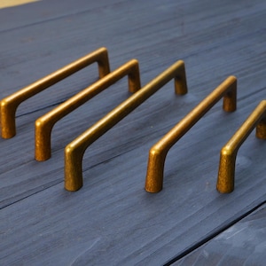 May include: Five brass-colored metal cabinet pulls arranged in a row, decreasing in length from left to right. Each handle features a smooth, arched design with two vertical mounting posts and a textured, slightly weathered metallic finish.