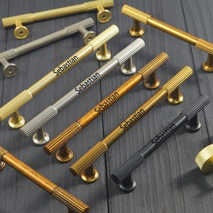 May include: A collection of metal cabinet pulls and knobs in various finishes including gold, silver, brass, and matte black. Each pull features a central smooth bar with ribbed ends and a pedestal base. The word Sebastian is printed on several pulls.