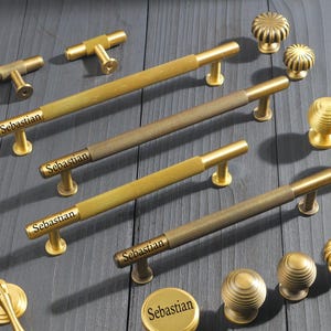 May include: A collection of brass-toned cabinet hardware including long bar pulls with knurled textures, round knobs with ribbed details, and drop pulls. Each piece features the word Sebastian printed in black on the metal surface.
