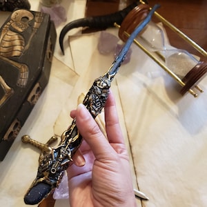 May include: Ornate, dark-colored wand with gold and silver accents. The wand features a detailed handle with a sculpted face and intricate designs. The tip has a dark, sparkling stone. The wand is held in a hand, with other mystical items in the background.