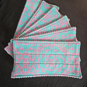 May include: A set of six teal and pink patterned reusable face cloths. The cloths are arranged in a fan shape, showcasing the floral design. The edges are finished with a white trim. The background is black.