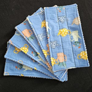 May include: A stack of blue, quilted fabric squares with a repeating pattern of watering cans in yellow, peach, and white. The squares have a white, textured border and are arranged in a fan shape.