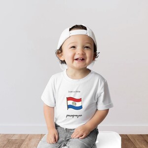 Puede incluir: Una camiseta blanca con la bandera paraguaya y la frase "Just a little paraguayan". La camiseta la lleva un niño con una gorra de béisbol blanca y vaqueros azules. El niño está sonriendo.