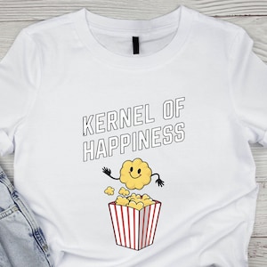 Puede incluir: Camiseta blanca con el texto "KERNEL OF HAPPINESS" en letras negras delineadas. Un personaje de palomitas de dibujos animados con una cara sonriente y brazos está encima de una caja de palomitas a rayas rojas y blancas.