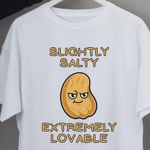 May include: White t-shirt with a cartoon peanut illustration. The text above the peanut reads "SLIGHTLY SALTY", and below it says "EXTREMELY LOVABLE". The peanut has a grumpy expression.