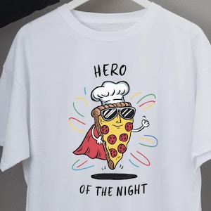 May include: White t-shirt featuring a cartoon pizza slice wearing sunglasses, a chef's hat, and a red cape. The text "HERO OF THE NIGHT" is printed above and below the pizza illustration. The shirt is hanging on a white hanger.